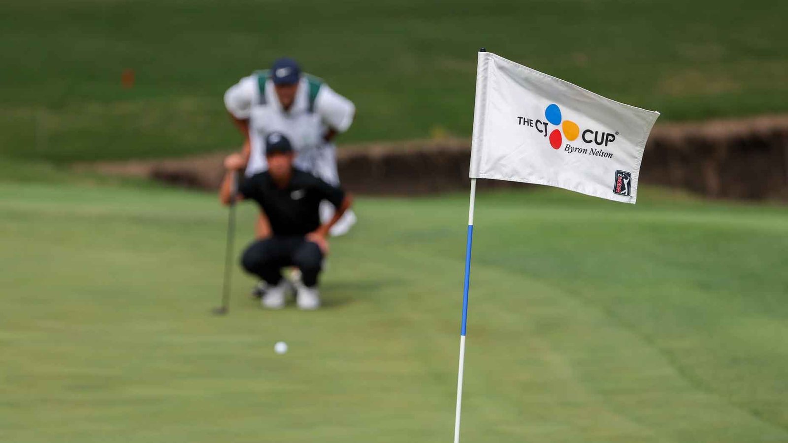 The Byron Nelson CJ Cup flag is shown on the 121st green with Doug Ghim (USA) in the background puttingt during the first round of the PGA CJ Cup Byron Nelson on May 2, 2024, TUP Craig Ranch in McKinney, TX.